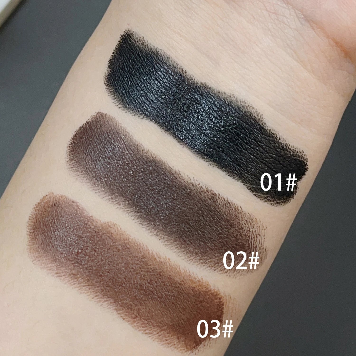 Three swatches of eyeshadow on a person's arm with labels indicating shades 01#, 02#, and 03#. The swatches are black, brown, and a darker brown.